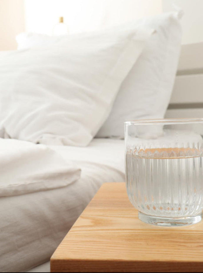 Clear glass with water on a wooden nightstand next to a bed with white bedding.
